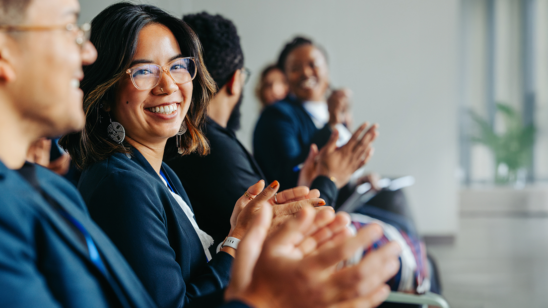 Group of diverse professionals sitting in a row, smiling and clapping at a conference or meeting.
